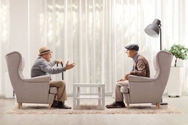 Koltuklarda oturmuş, bir odada sohbet eden iki yaşlı adam.