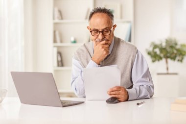 Düşünceli, olgun bir adam dizüstü bilgisayarın önünde oturuyor ve kağıt bir belgeye bakıyor.