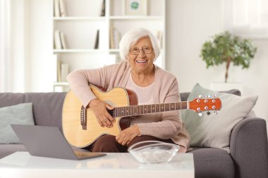 Evde bir kanepede oturan yaşlı bir kadın. Akustik gitarı ve masada dizüstü bilgisayarı var.