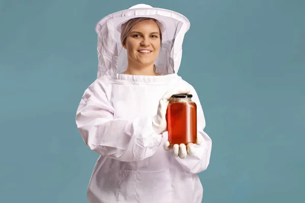 身穿制服的年轻女性养蜂人 手里拿着一罐蓝色背景的蜂蜜 — 图库照片