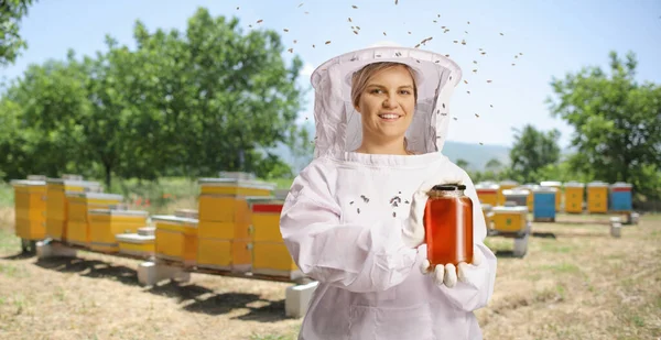 Young female bee keeper in a uniform holding a jar of honey on apiary and bees flying