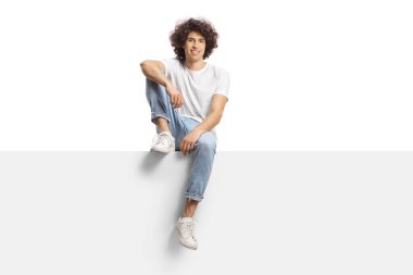 Casual young man with curly hair sitting on a blank panel isolated on white background