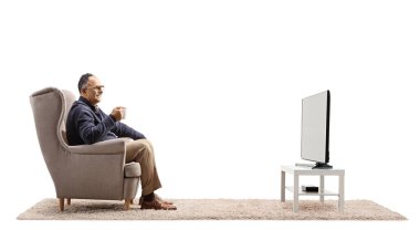 Profile shot of a mature man sitting in an armchair watching tv and holding a cup isolated on white background