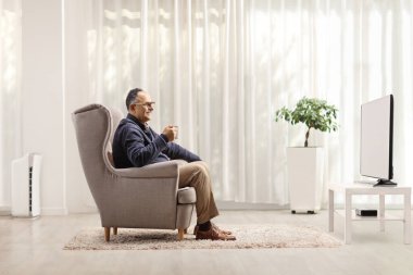 Olgun bir adamın oturduğu koltukta oturup televizyon izlediği ve oturma odasında elinde bardak tuttuğu profil fotoğrafı.