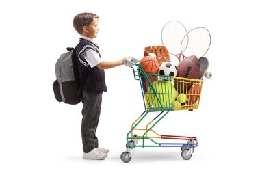 Üniformalı bir çocuğun beyaz arka planda izole edilmiş spor malzemeleriyle dolu bir alışveriş arabasıyla tam boy profil fotoğrafı.