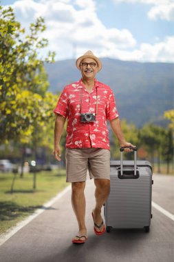 Gülümseyen olgun bir erkek gezgin bavulu çekiyor ve asfalt bir yolda yürüyor.