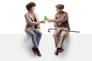 Neşeli genç adam boş bir panelde oturuyor ve beyaz arka planda izole edilmiş yaşlı bir adamla bira şişeleriyle kadeh kaldırıyor.
