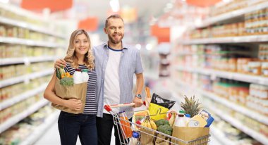 Young couple with a shopping cart full of food and a grocery bag inside a supermarket