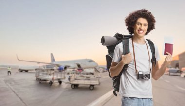 Young male tourist with a backpack showing a passport and smiling at an airport apron