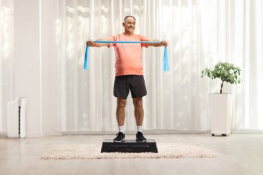 Mature man in sportswear exercising with a stretch strap on a step aerobic platform inside a room