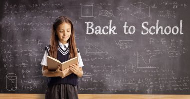 Schoolgirl reading a book in front of a blackboard with math formulas and text back to school
