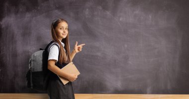 Schoolgirl with a backpack and book pointing at a blackboard and looking at camera
