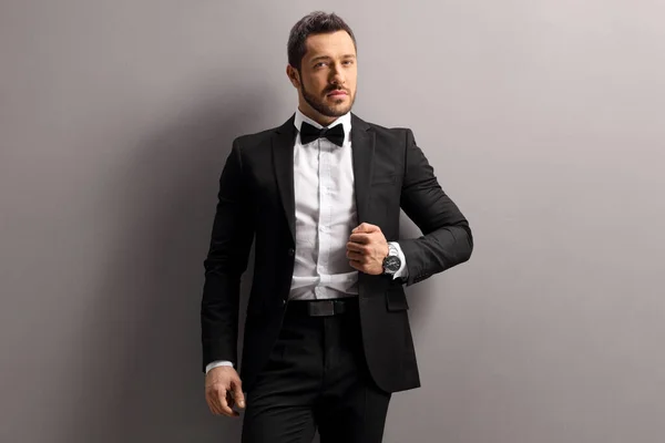 Male model in a black suit and bow tie leaning on a gray wall