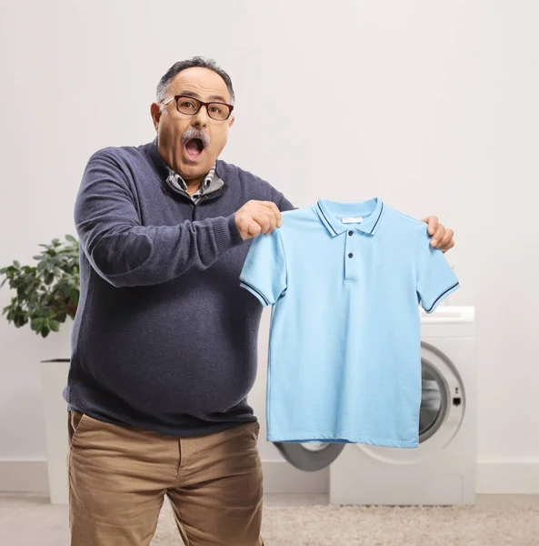 Confused Mature Man Holding Shrunken Shirt Front Washing Machine — Stockfoto