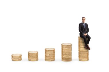 Professional man in suit and tie sitting on a rising piles of coins and looking at camera isolated on white background