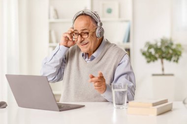 Mature man sitting at a table listening with headphones and looking at a laptop computer at home