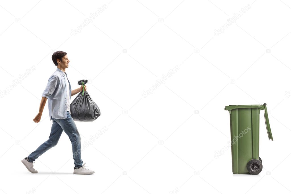 Full length profile shot of a casual young man walking towards a bin and carrying a plastic garbage bag isolated on white background