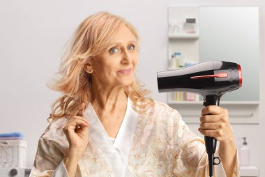 Banyoda saç kurutma makinesi kullanan bornozlu güzel olgun bir kadın.