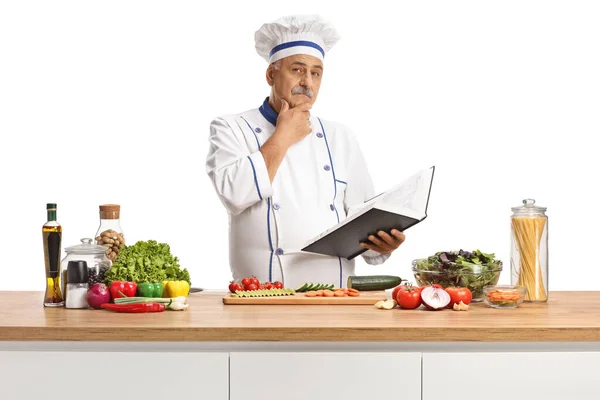 成熟的男性厨师在厨房柜台后面拿着一本烹饪书 在白色背景下独立思考 — 图库照片