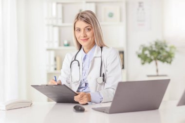 Genç bir bayan doktor ofiste oturuyor ve bir masada dizüstü bilgisayarla bir belge yazıyor.