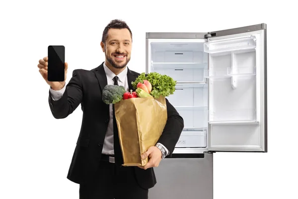 Businessman Holding Bag Groceries Showing Smartphone Front Empty Fridge Isolated — Stock Photo, Image