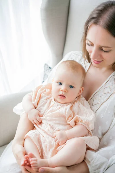 Happy young mother holds her little daughter 5 months old in her arms sitting in a chair at home.