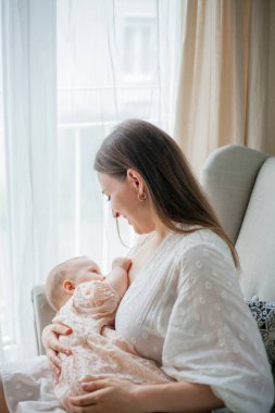 Beyaz elbiseli güzel bir anne, 5 aylık kızını emziriyor. Pencerenin yanındaki sandalyede oturuyor..