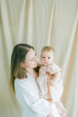 Beyaz elbiseli güzel bir anne, 5 aylık küçük mavi gözlü kızını kollarında tutuyor..