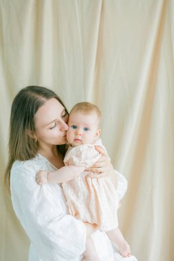 Beyaz elbiseli güzel bir anne, 5 aylık küçük mavi gözlü kızını kollarında tutuyor..