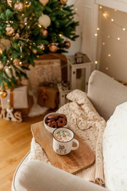 Noel süsleriyle dolu oturma odası. Bir fincan kremalı sıcak kakao, ahşap tepside kurabiyeler. Noel ağacı, hediye kutuları, rahat koltuk, yastıklar, şömine..