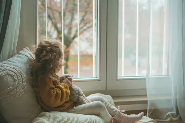 Pequeña linda chica de pelo rizado en jersey de punto se sienta delante de la ventana. Acogedora casa. Otoño cálido. — Foto de Stock