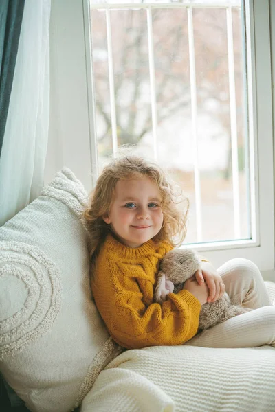 Piccola graziosa ragazza dai capelli ricci in pullover lavorato a maglia si siede davanti alla finestra. Casa accogliente. Autunno caldo. — Foto Stock