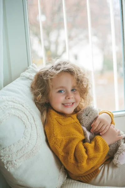 Piccola graziosa ragazza dai capelli ricci in pullover lavorato a maglia si siede davanti alla finestra. Casa accogliente. Autunno caldo. — Foto Stock