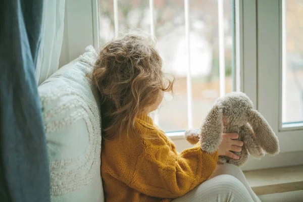 Pequeña linda chica de pelo rizado en jersey de punto se sienta delante de la ventana. Acogedora casa. Otoño cálido. — Foto de Stock