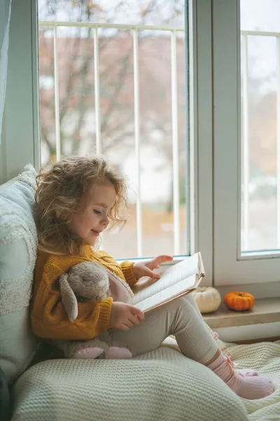 A little cute curly haired girl in knitted pullover is reading a book in front of the window. Cozy home. Warm autumn. Mood. — Stock Photo, Image
