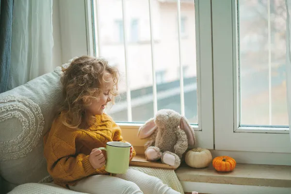 Pequena menina encaracolado bonito em pulôver de malha é beber chá e comer biscoitos na frente da janela. Uma casa acolhedora. Outono quente. — Fotografia de Stock