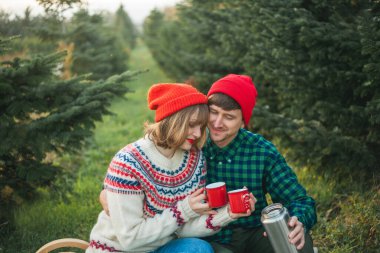 Noel zamanı. Kırmızı şapkalı ve kazaklı mutlu genç çift Noel ağaçlarının yakınındaki termostan sıcak çay içerler. Noel ağacı pazarı.