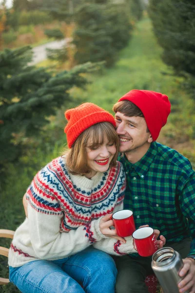 Kerstmis. Gelukkig jong koppel verliefd in rode hoeden en truien drinken warme thee uit thermoskan in de buurt van kerstbomen. Kerstboommarkt. — Stockfoto