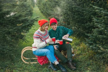 Noel zamanı. Kırmızı şapkalı ve kazaklı mutlu genç çift Noel ağaçlarının yakınındaki termostan sıcak çay içerler. Noel ağacı pazarı.