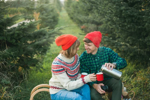 Święta. Szczęśliwa młoda para zakochana w czerwonych kapeluszach i swetrach pije gorącą herbatę z termosu w pobliżu choinek. Rynek choinek. — Zdjęcie stockowe