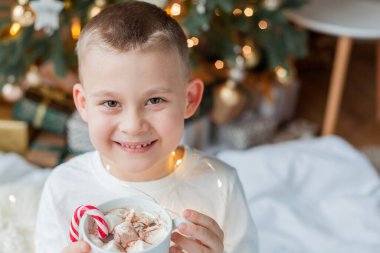 Şenlikli pijamalı 6 yaşındaki sevimli çocuk Noel ağacının yanında elinde noel süslemelerinde lolipop ve lokum dolu bir kupa ile oturuyor. Kış tatili Yeni Yıl.