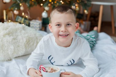 Şenlikli pijamalı 6 yaşındaki sevimli çocuk Noel ağacının yanında elinde noel süslemelerinde lolipop ve lokum dolu bir kupa ile oturuyor. Kış tatili Yeni Yıl.
