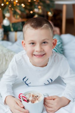 Şenlikli pijamalı 6 yaşındaki sevimli çocuk Noel ağacının yanında elinde noel süslemelerinde lolipop ve lokum dolu bir kupa ile oturuyor. Kış tatili Yeni Yıl.