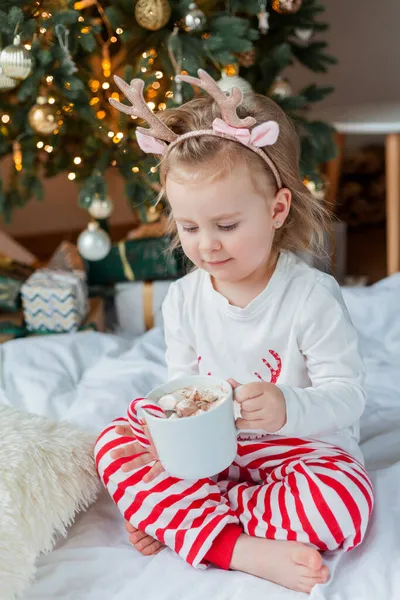 穿着节日睡衣的可爱的2岁女孩坐在圣诞树旁 手里拿着一大杯热可可 上面装饰着棉花糖和棒棒糖 寒假农历新年 — 图库照片