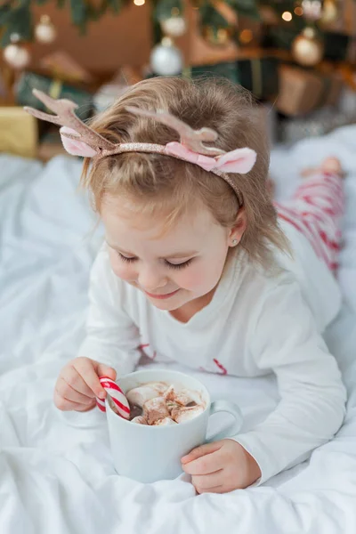 穿着节日睡衣的可爱的2岁女孩坐在圣诞树旁 手里拿着一大杯热可可 上面装饰着棉花糖和棒棒糖 寒假农历新年 — 图库照片