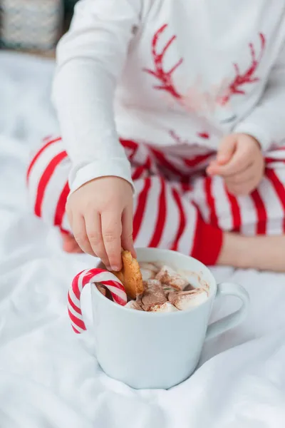 お祝いのパジャマの愛らしい2歳の女の子は クリスマスの装飾でマシュマロとロリポップとホットココアのマグカップを持ってクリスマスツリーに座っています 冬休みだ — ストック写真