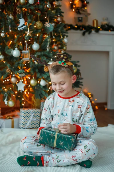 穿着睡衣的可爱的7岁男孩在圣诞树旁打开礼品盒 新年的灯 寒假舒适的家 — 图库照片