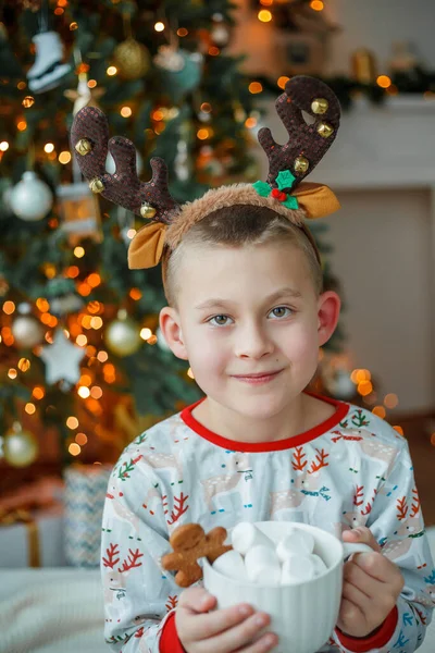 可爱的穿着睡衣的7岁男孩在圣诞树前喝着热可可 还喝着棉花糖和姜饼饼干 新年的灯 寒假舒适的家 — 图库照片