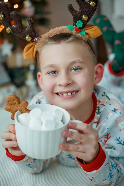 可爱的穿着睡衣的7岁男孩在圣诞树前喝着热可可 还喝着棉花糖和姜饼饼干 新年的灯 寒假舒适的家 — 图库照片