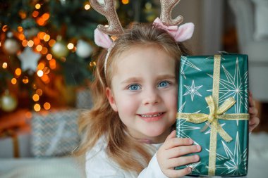 Pijamalı 3 yaşında sevimli bir kız Noel ağacının yanındaki hediye kutusunu açıyor. Yeni yıl ışıkları. Kış tatili Konforlu ev.
